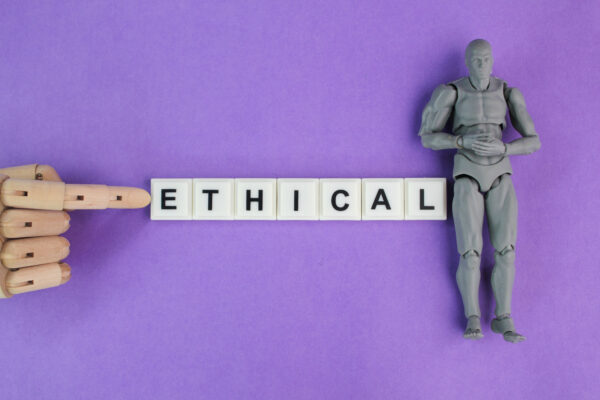 Ein Finger einer hölzernen Hand zeigt auf den Schriftzug Ethical, der aus einzelnen Buchstaben-Bausteinen zusammengesetzt ist. Rechts daneben ist eine graue Figur zu sehen, die ein Mensch oder ein Roboter sein könnte.