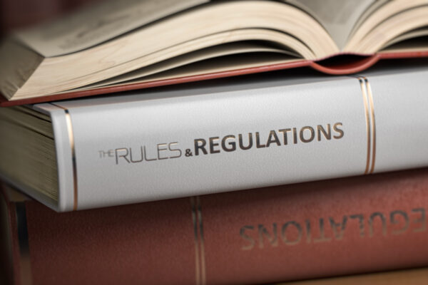Drei Bücher liegen übereinander, das dritte obenauf ist aufgeschlagen, Auf dem Buchrücken des mittleren Buchs ist die Beschriftung "Rules & Regulations" zu erkennen.