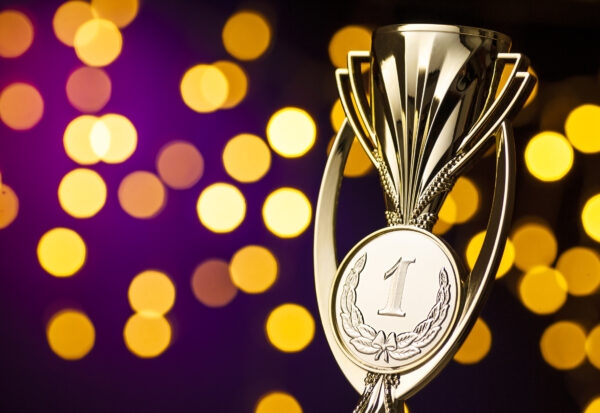 Im Vordergrund ist ein goldener Pokal zu sehen, der mit einer eins für den ersten Platz beschriftet ist. Der Hintergrund ist lila bis schwarz mt leuchtenden pailletten-artigen Highlights.