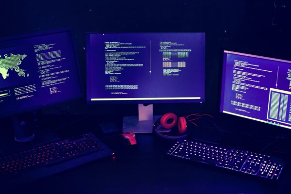 Drei Computerbildschirme stehen in einem abgedunkelten Raum, es leigen noch Kopfhörere und Tastatur bereit. Die Bildschirme zeigen eine textbasierte Computeranwendung.