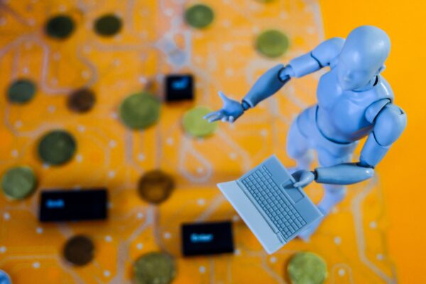 Ein humanoider Roboter, rechts im Bild zu sehen, hält einn weißen Laptop in einer Hand. Der Roboter scheint auf einem Untergrund zu stehen, der aus einer Platine besteht und einen gelben Hintergrund besitzt.
