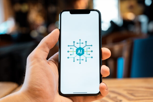 Eine Hand hält ein Smartphone auf dem symbolhaft eine AI-Anwendung dargestellt ist.
