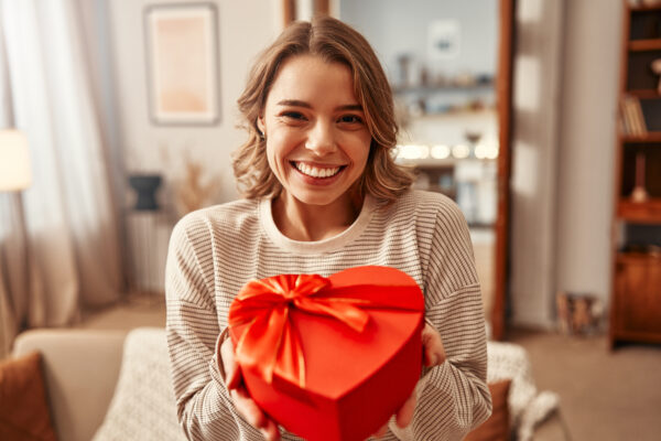 Eine lächelnde Frau hält eine herzförmige Geschenkschachtel in die Kamera.